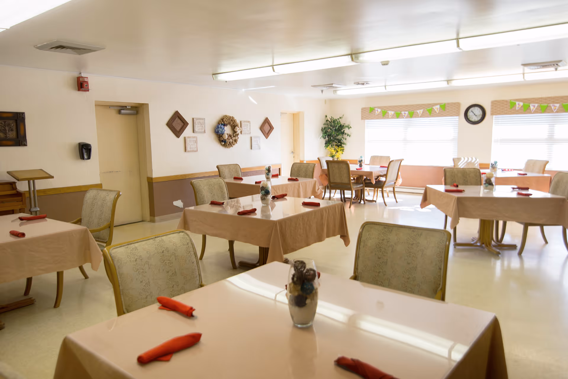A bright dining room with several tables covered in beige tablecloths, each set with red napkins and a small decorative centerpiece. The room has cushioned chairs around the tables, large windows with blinds letting in natural light, a clock on the wall, and some wall decorations including a wreath and framed art. A potted plant is placed near the windows.