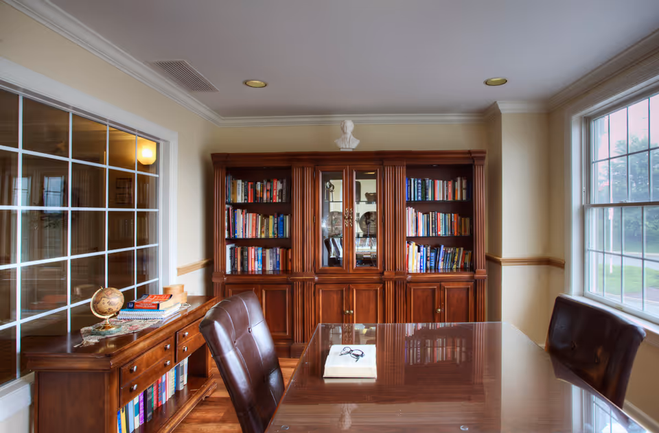 A cozy study or reading room with a large wooden bookshelf filled with books against the back wall. In front of the bookshelf is a polished wooden table with a book and a pair of glasses on it. Two brown leather chairs are positioned around the table. To the left, there is a wooden sideboard with more books and a small globe. Large windows on the right and a glass-paneled wall on the left allow natural light to fill the room.