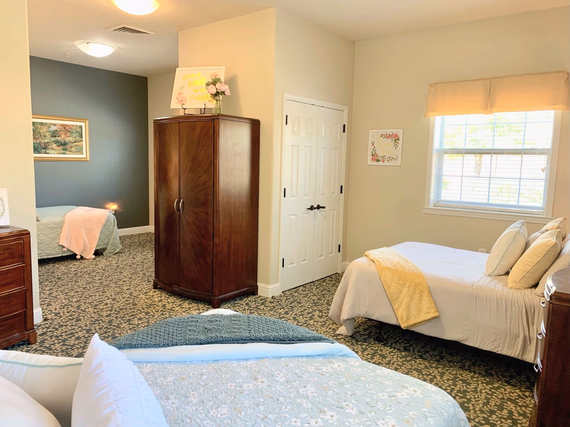 A bright and cozy bedroom in a senior living facility with two beds, one in the foreground with light blue bedding and another against the wall with white bedding and a yellow throw blanket. The room features patterned carpet, a wooden armoire, a dresser, a window with blinds and a beige valance, and framed artwork on the walls.