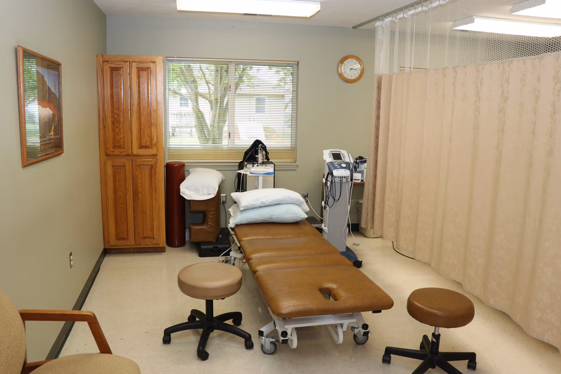 A medical examination room with a brown exam table, rolling stools, medical equipment, a privacy curtain, and a window.