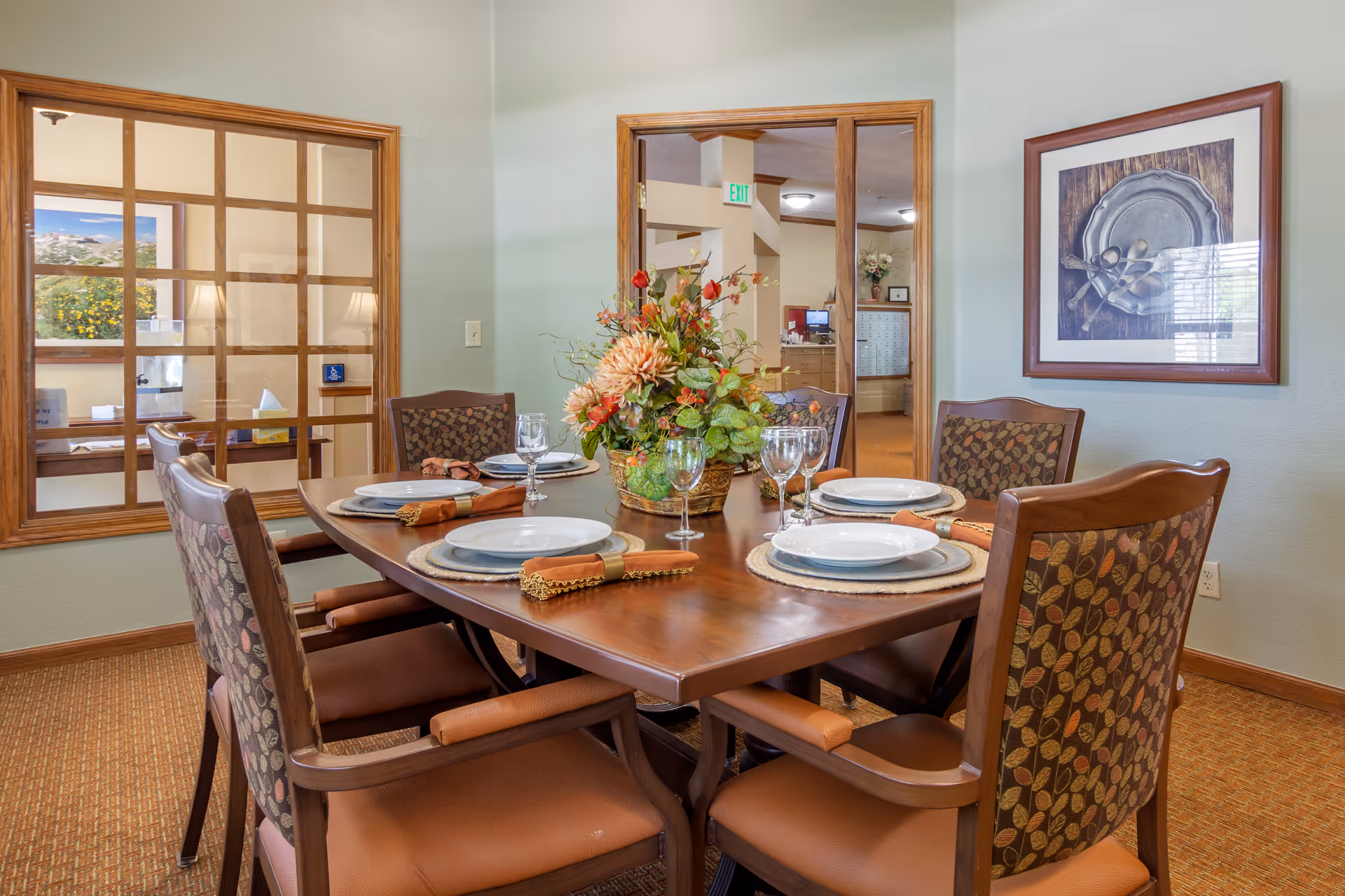A dining room with a wooden table set for six people, featuring white plates, wine glasses, and brown napkins with gold trim. A floral centerpiece decorates the middle of the table. The room has light green walls, a framed picture of silverware on the wall, and a window with wooden grid framing looking into another room.