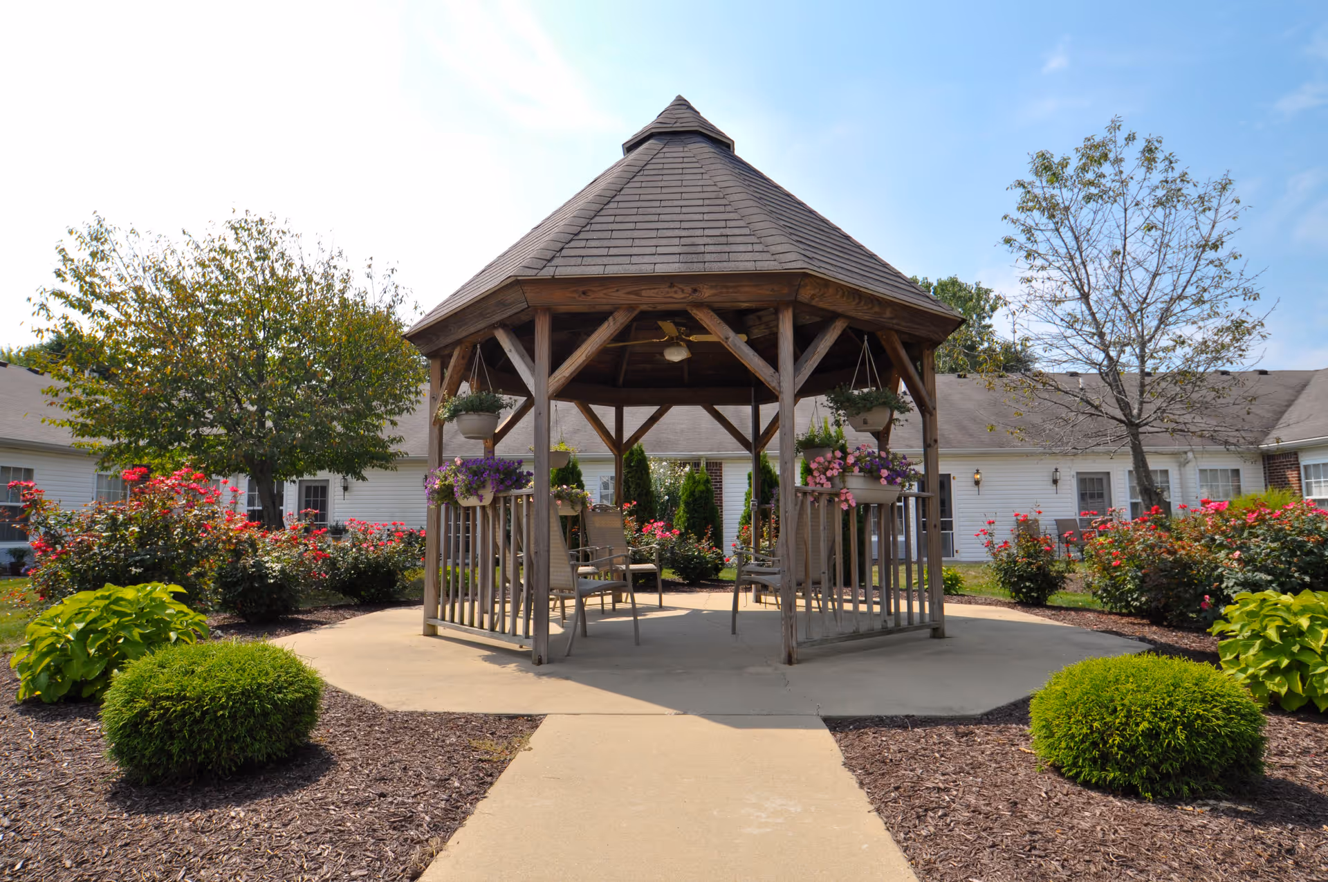 A wooden gazebo with a shingled roof is situated in a garden area with various green shrubs and blooming flowers. The gazebo contains several chairs and hanging flower pots. Surrounding the garden are single-story white buildings with windows and doors. The sky is clear and blue.