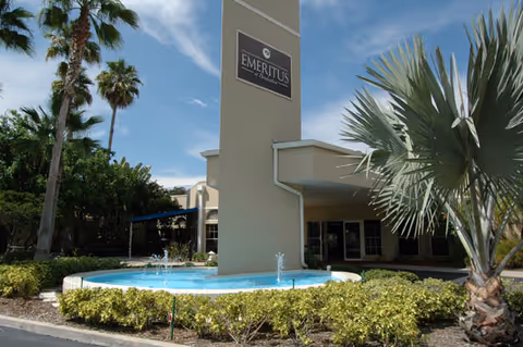 Front entrance of a senior living facility with a tower sign, fountain, palm trees, and landscaped shrubs.