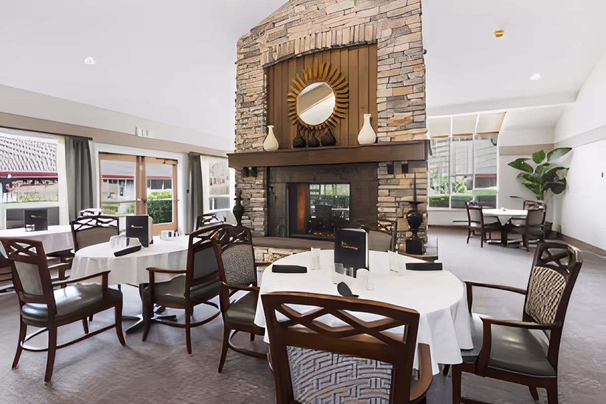 A dining room with round tables covered in white tablecloths and surrounded by wooden chairs with cushioned seats. In the center of the room is a large stone fireplace with a decorative sunburst mirror and vases on the mantel. Large windows and glass doors allow natural light to fill the space, and there is a large green plant in the corner.