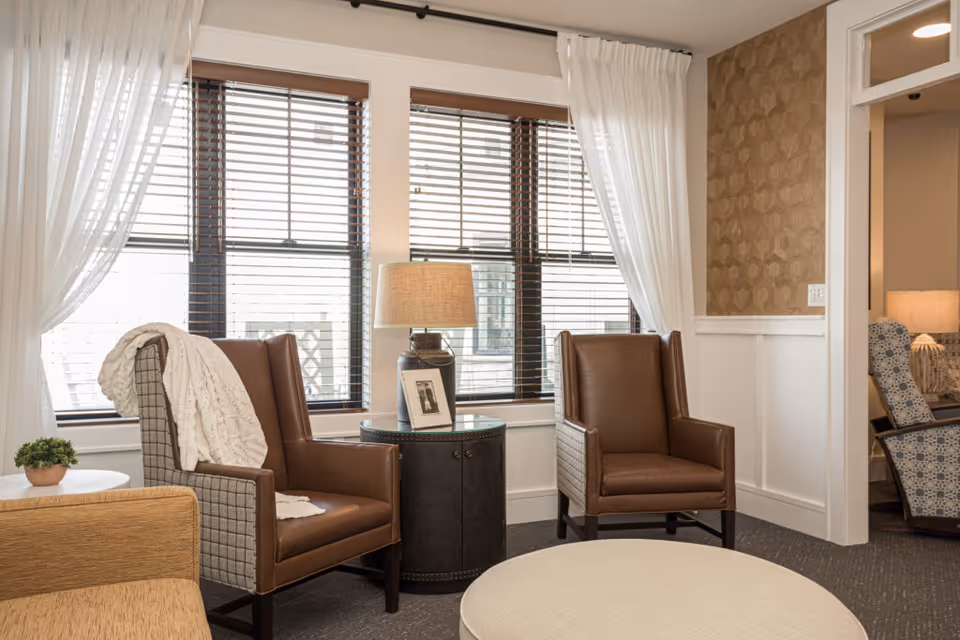 A cozy sitting area with two brown leather armchairs, a round side table with a lamp and framed photo, a small potted plant on a white table, and large windows with wooden blinds and sheer white curtains. The room has neutral tones and patterned wallpaper on one wall.