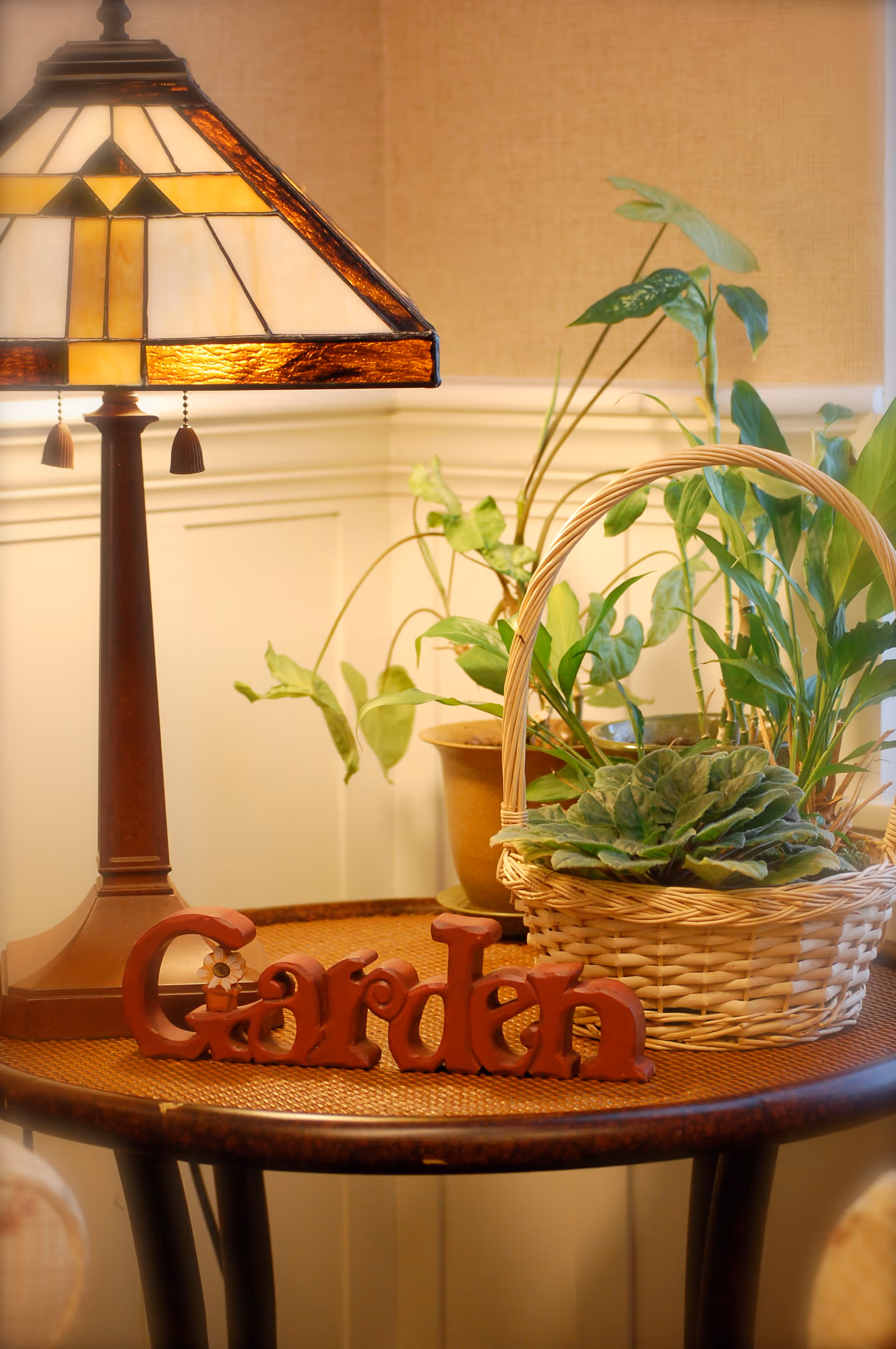 A close-up of a round wooden table with a stained glass lamp, a basket with green plants, a potted plant, and a decorative wooden sign that reads 'Garden'. The background shows a beige wall with white wainscoting.