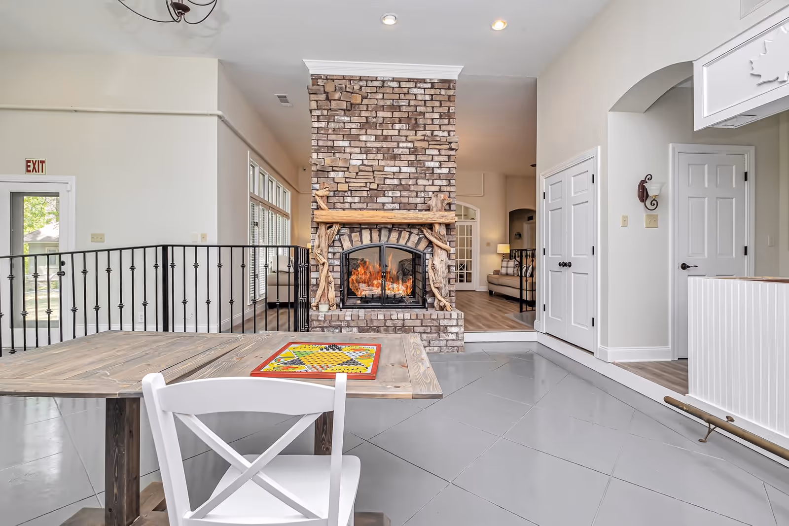 Bright communal living room with a central brick fireplace, a wooden table and white chair in the foreground, and a seating area beyond.
