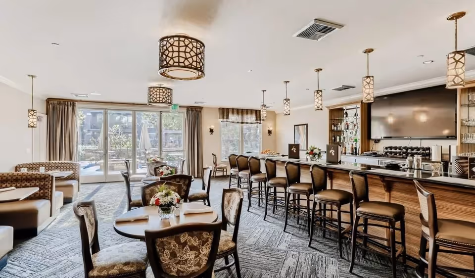 Bright upscale dining and lounge area with tables, patterned chairs, a long bar with stools, pendant lights, and large windows looking out to a patio.