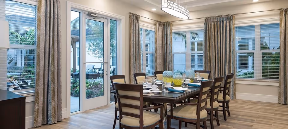 Bright dining room with a large wooden table set for eight, cushioned chairs, floor-to-ceiling windows and glass doors opening to an outdoor patio.