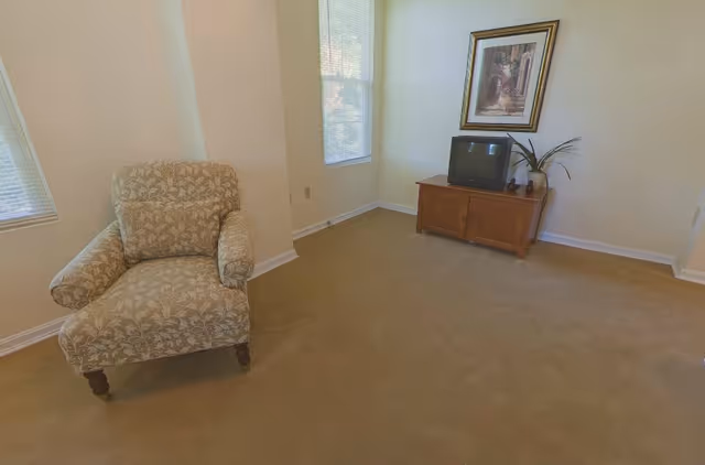 A sparsely furnished living room with a patterned armchair, a small TV on a wooden cabinet, framed artwork, and windows.