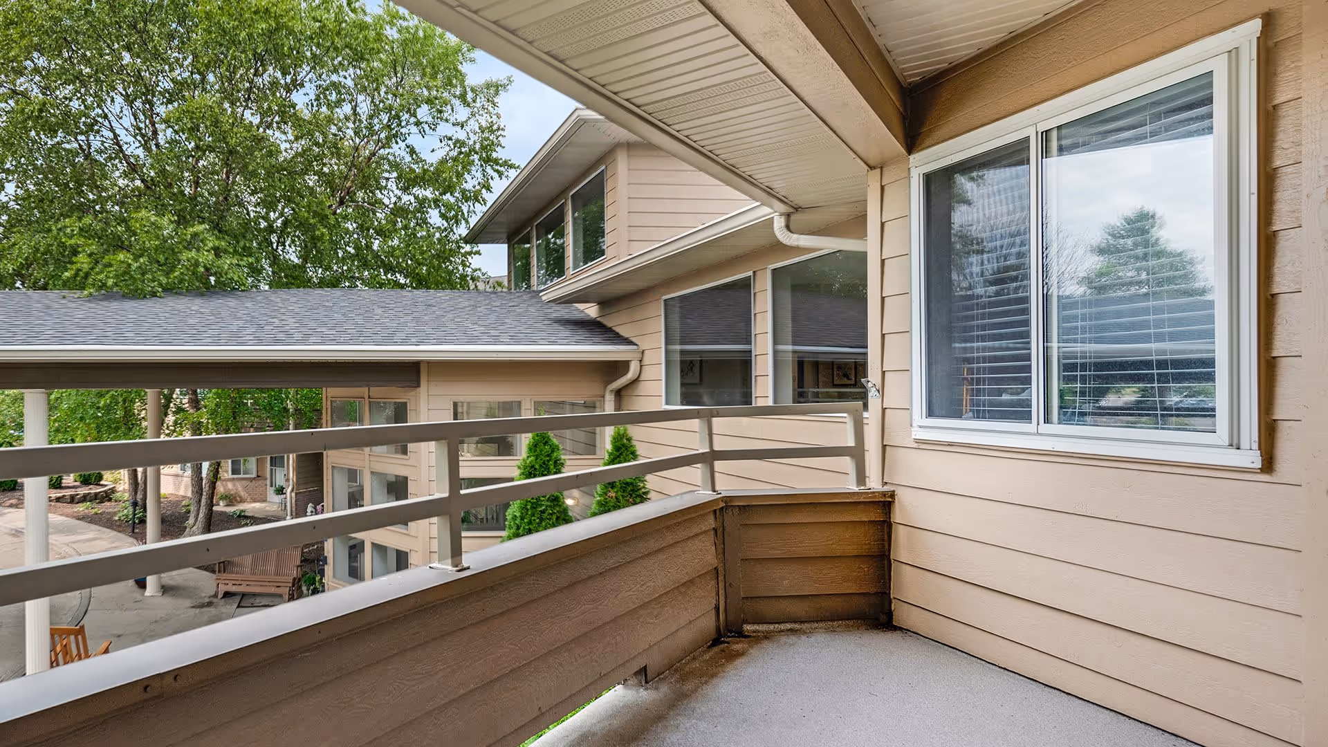 View of a covered balcony area with beige siding and railing, overlooking a courtyard with trees and benches at Holiday Palmer Hills facility.