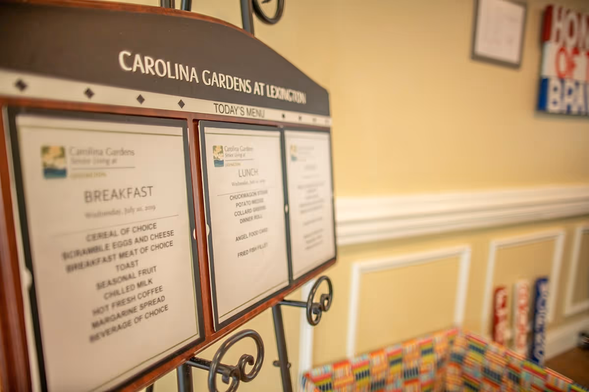 A menu board displaying breakfast and lunch options at Carolina Gardens at Lexington, placed indoors against a yellow wall with white trim. The menu includes items like cereal, scrambled eggs, potato wedges, and fried fish fillet. There is a colorful basket and some signs in the background.