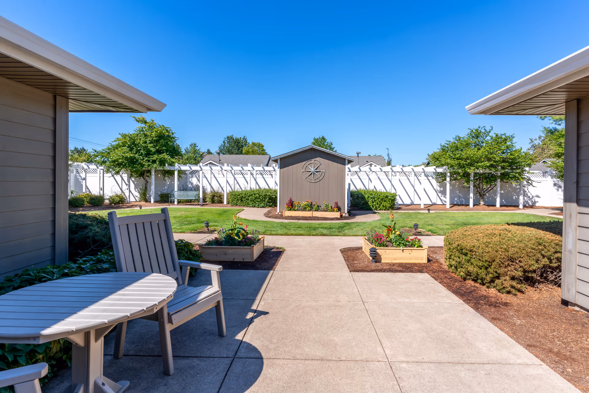 Sunny fenced courtyard with patio seating, raised flower beds, a small shed, and landscaped lawn.