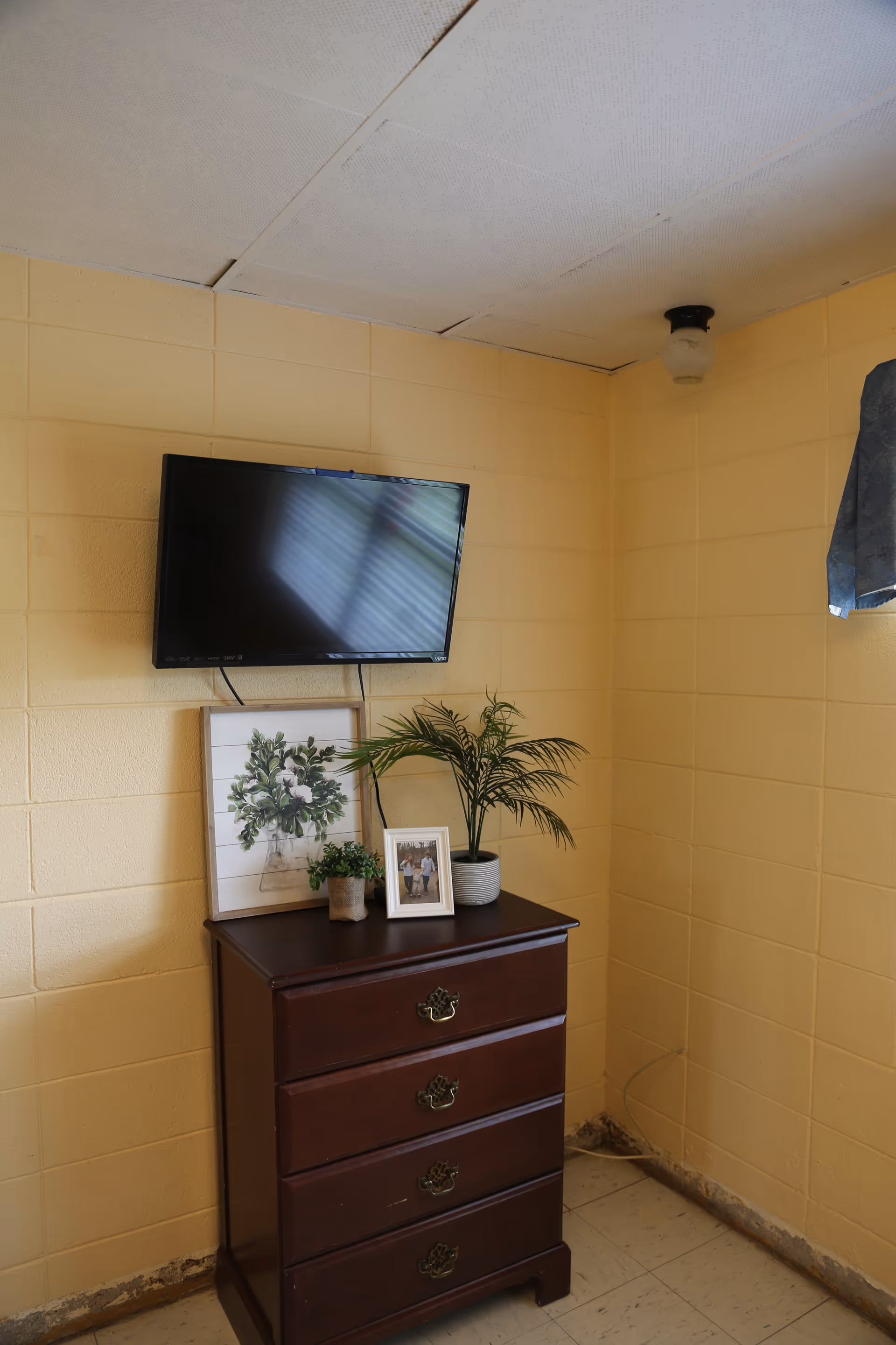 A corner of a room with yellow painted cinder block walls and a white tiled floor. A dark wooden chest of drawers with four drawers is placed against the wall. On top of the chest are three potted plants and a framed photo. Above the chest, a flat-screen TV is mounted on the wall. The ceiling has white tiles, some of which appear to be slightly misaligned. A small window with a blue curtain is partially visible on the right side.