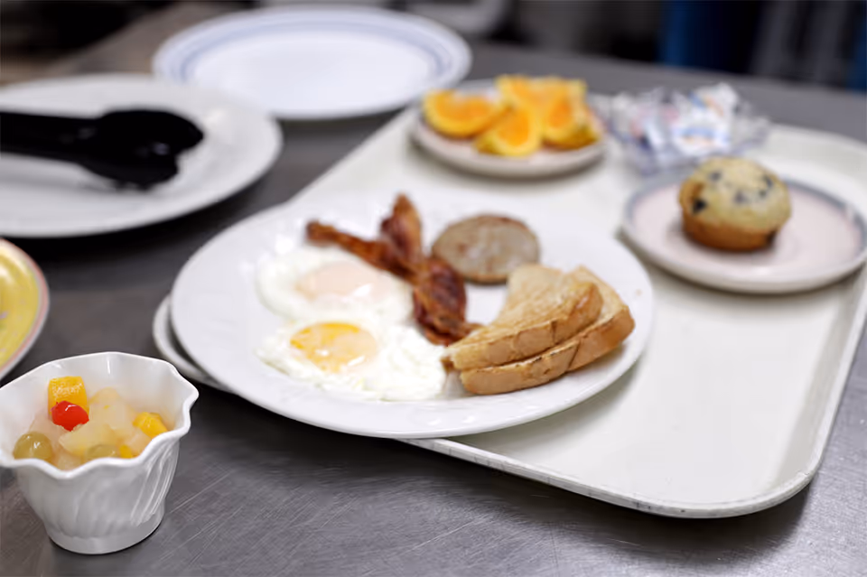 Cafeteria breakfast tray with fried eggs, bacon, toast, a sausage patty, a blueberry muffin and a cup of mixed fruit.