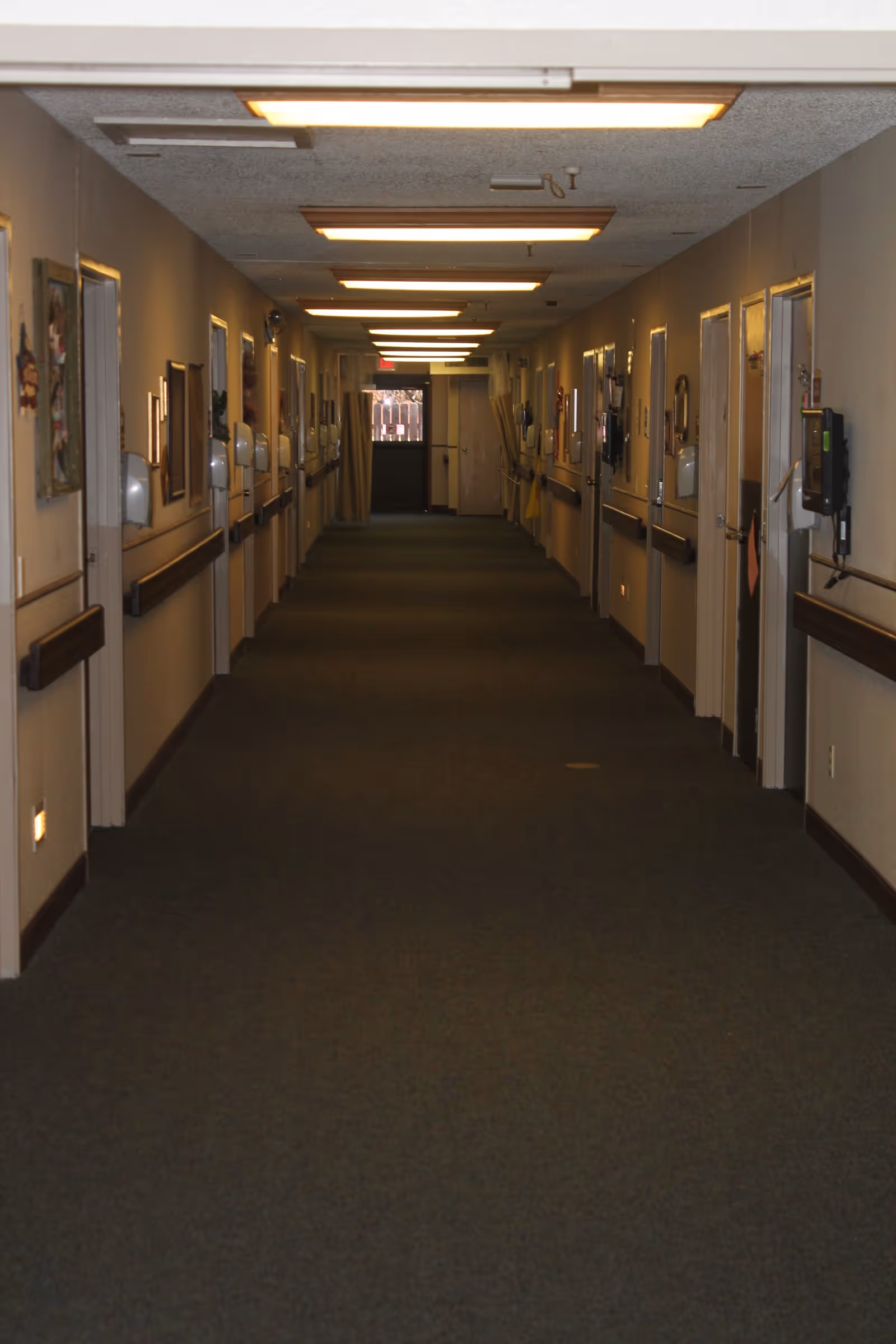 A long, carpeted hallway in a senior living facility with multiple closed doors on both sides. The walls are decorated with framed pictures and there are handrails along the length of the corridor. Ceiling lights illuminate the hallway, and there is an exit door at the far end.