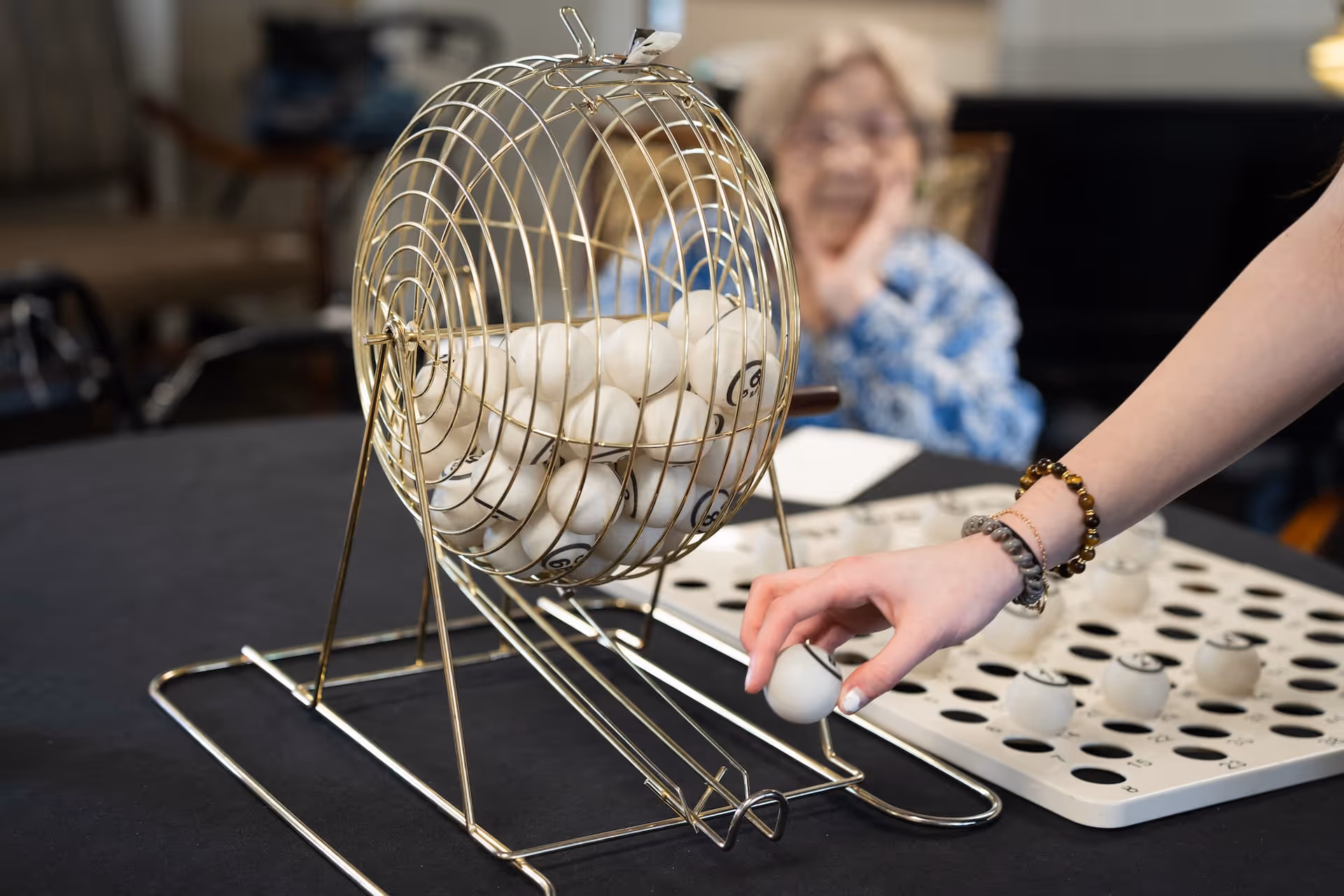 A close-up of a bingo cage filled with numbered balls on a table, with a person's hand reaching to pick a ball. In the background, an elderly woman is sitting and watching the game.