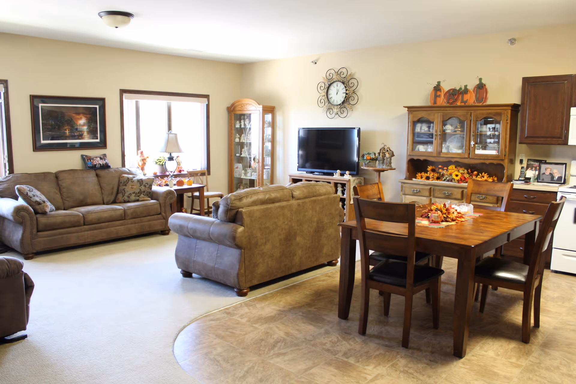 A cozy living and dining area in a senior living facility with two brown sofas, a wooden dining table with four chairs, a TV on a stand, a wall clock, a glass display cabinet, and kitchen cabinets. The room is decorated with autumn-themed items and has large windows letting in natural light.