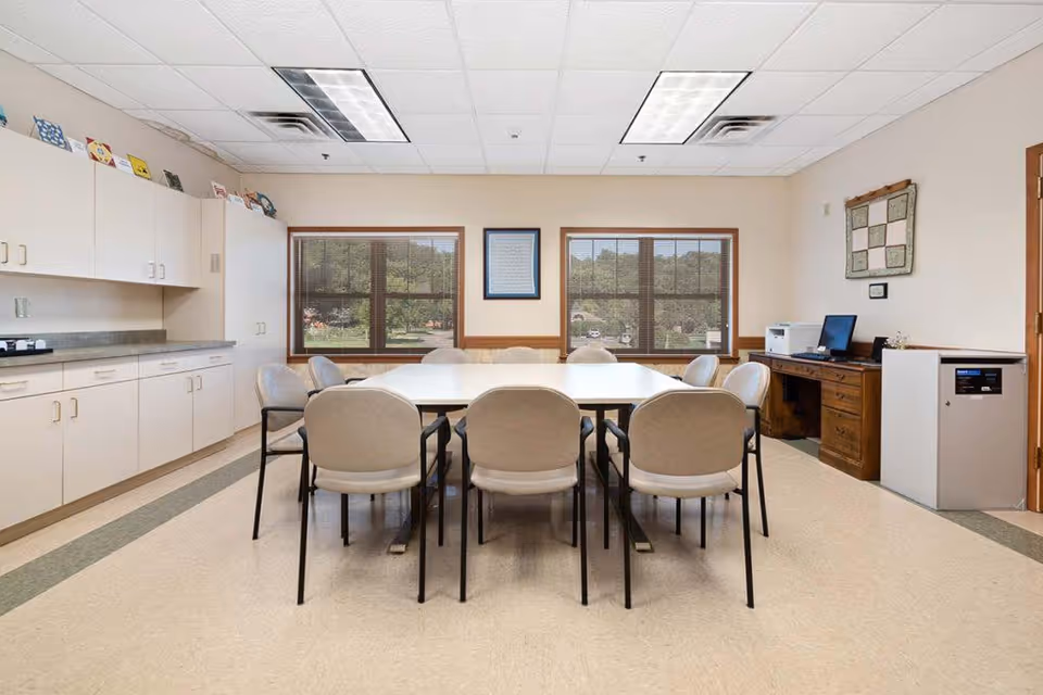 A meeting or activity room with a large rectangular table surrounded by eight beige chairs. The room has two large windows with views of greenery outside. On the left side, there are white cabinets and a countertop. On the right side, there is a wooden desk with a computer, printer, and a small white appliance next to it. The walls are light-colored with a framed picture and a quilt hanging.