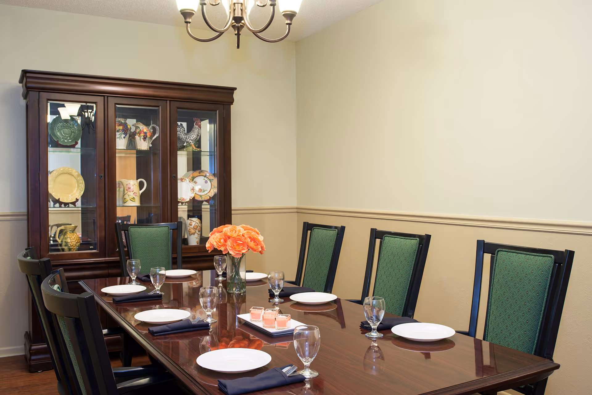 A formal dining room with a polished wooden table set for six people. The table has white plates, glassware, and navy blue napkins. A vase with orange flowers is placed in the center. Behind the table is a dark wooden china cabinet displaying decorative plates and pitchers. The walls are painted beige with a chair rail molding, and a chandelier hangs from the ceiling.