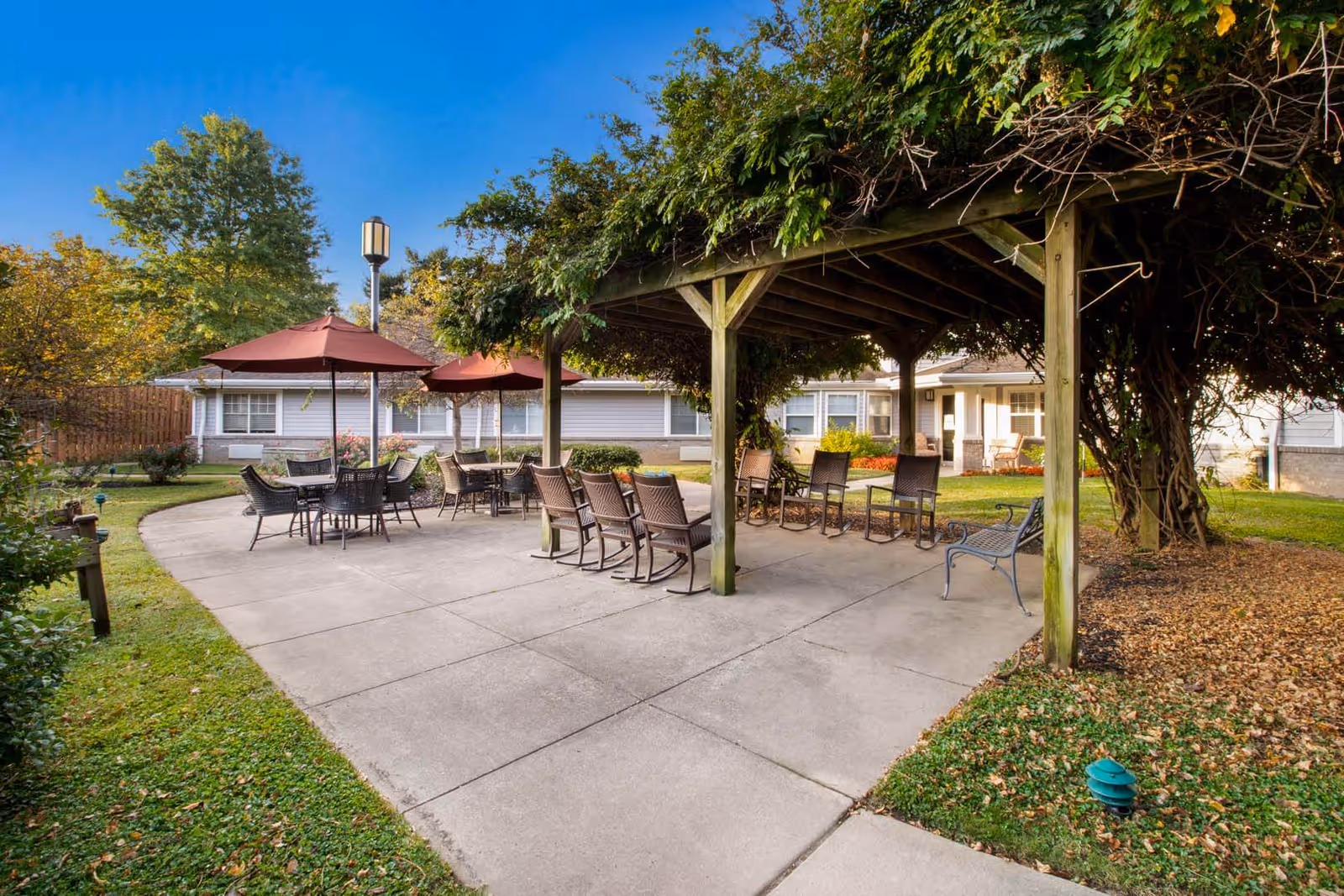 Outdoor patio area at Vitality Living Springdale featuring a wooden pergola covered with greenery, several chairs arranged under the pergola, and multiple tables with umbrellas on a concrete patio surrounded by grass and trees.