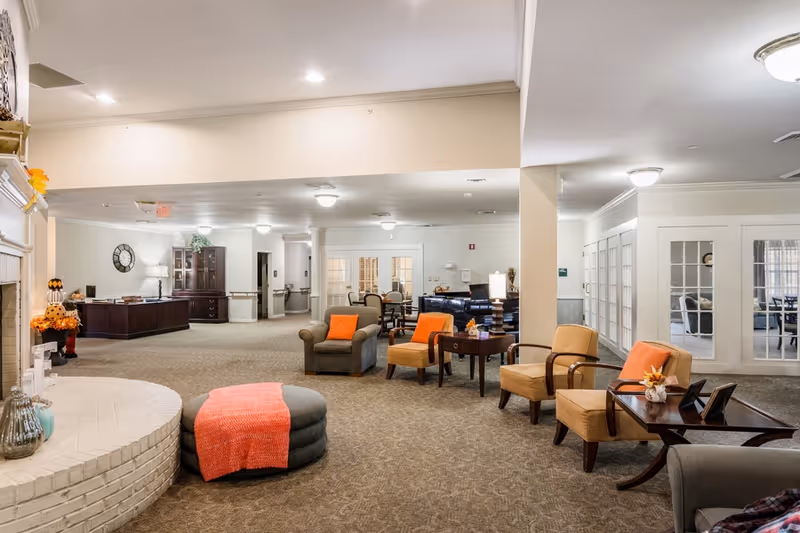 Spacious senior living facility common lounge with multiple armchairs, an ottoman draped with an orange throw, side tables, and a fireplace.