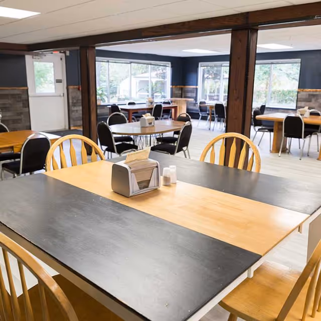 A spacious dining room with multiple wooden tables and chairs arranged neatly. The tables have napkin holders and salt and pepper shakers. Large windows allow natural light to brighten the room, and the walls have a combination of dark and light colors with wooden accents.