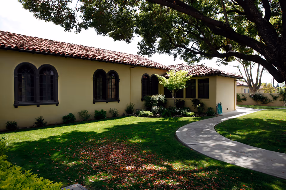 Single-story Mediterranean-style building with arched windows, a red tile roof, a curved concrete walkway, and a shaded lawn.