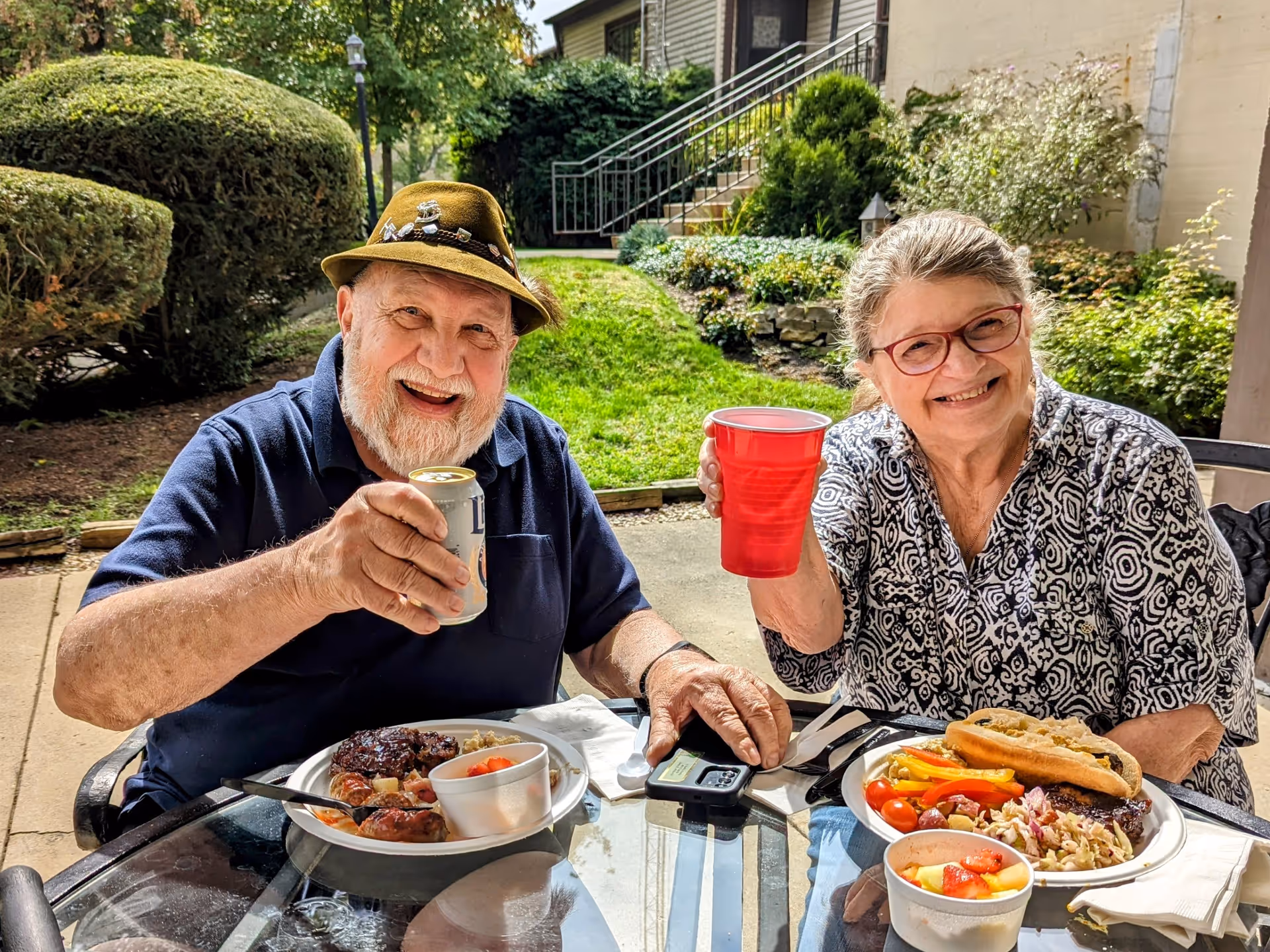 An elderly man and woman sitting at an outdoor glass table enjoying a meal together. The man is wearing a green hat and navy blue shirt, holding up a can of beer and smiling. The woman is wearing glasses and a patterned shirt, holding up a red plastic cup and smiling. Their plates have a variety of food including grilled meat, vegetables, pasta salad, and fruit cups. Behind them is a green lawn, bushes, and a building with stairs.