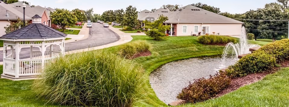 A landscaped outdoor area at a senior living facility featuring a small pond with a fountain, green grass, bushes, and a white gazebo. Residential buildings and a paved road with parked cars are visible in the background under a partly cloudy sky.