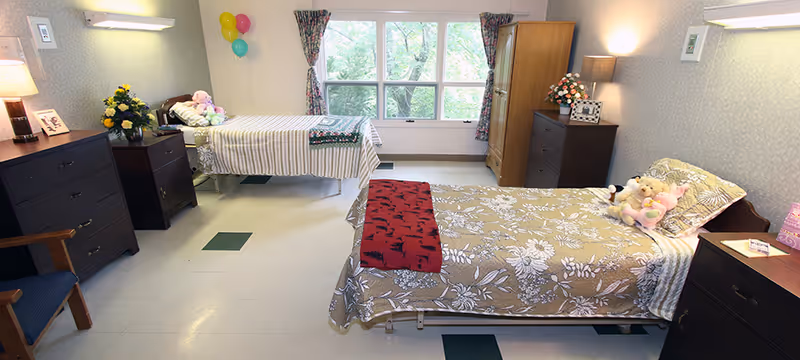 Shared senior living bedroom with two single beds, dressers, a large window, and decorative items.