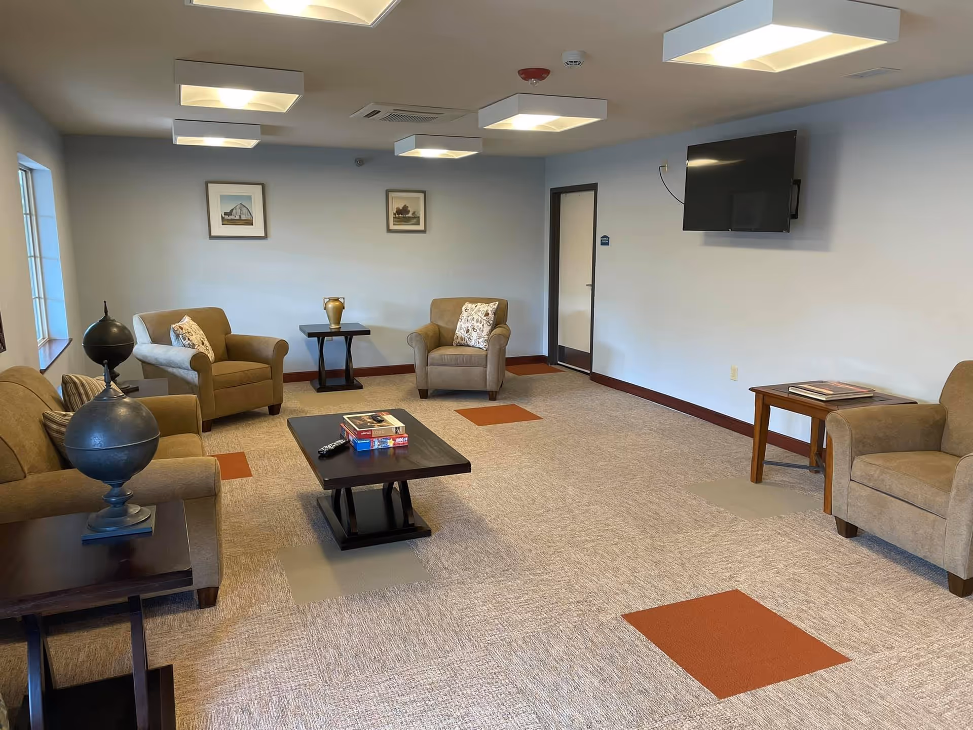 A cozy living room with beige armchairs arranged around a dark wooden coffee table with books and a remote control on it. There are two side tables, one with a decorative globe and the other with a vase. The room has light-colored walls with two framed pictures, a wall-mounted flat-screen TV, and ceiling lights. The carpet is light with orange and gray square patterns.