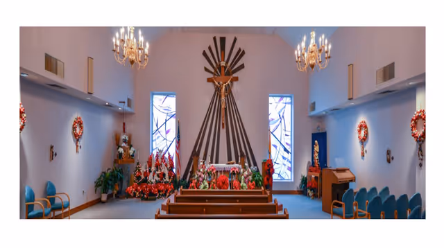 Interior view of a chapel decorated for Christmas with wreaths on the walls, poinsettias around the altar, stained glass windows, wooden pews, chandeliers, and a crucifix mounted on a wooden design behind the altar.
