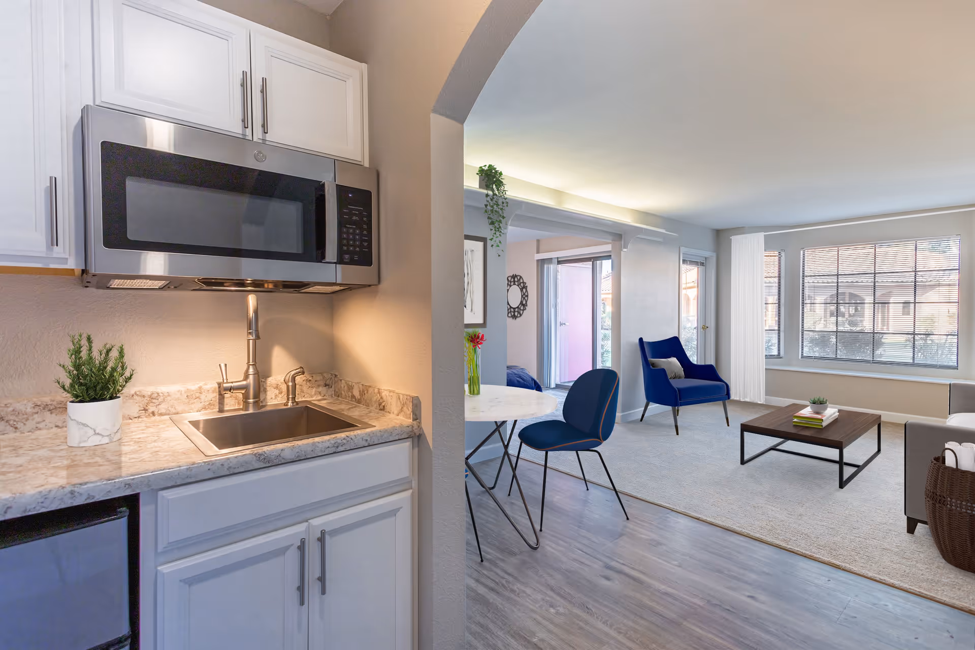 Interior view of a senior living facility showing a small kitchen area with a sink, countertop, and microwave on the left. The kitchen opens into a living room with a blue armchair, a coffee table, a dining table with two chairs, and large windows letting in natural light.