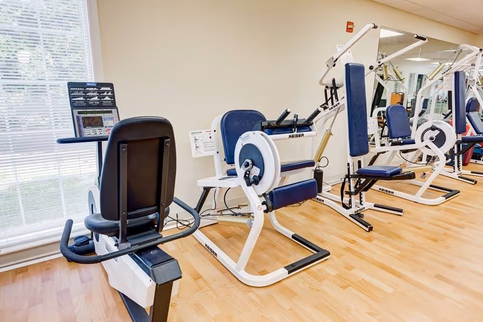A fitness room with exercise equipment including a recumbent bike and multiple strength training machines with blue padding, light wood flooring, a large window with blinds, and a mirrored wall reflecting the equipment.