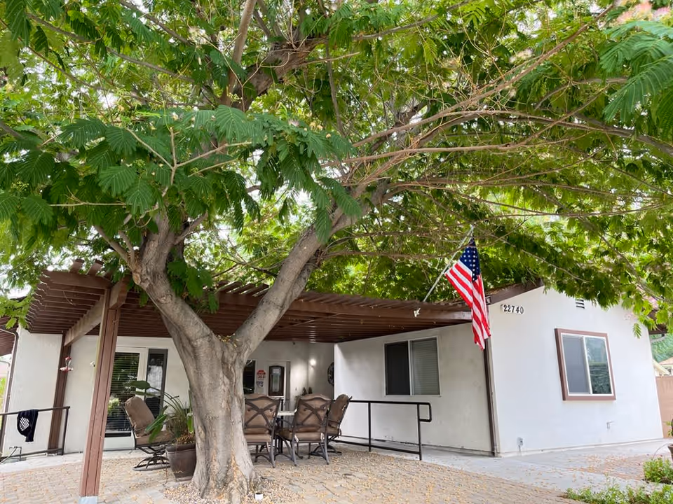 Outdoor patio area of a residential building with a large tree providing shade. There is a wooden pergola with outdoor chairs and a table underneath. An American flag is mounted on the corner of the building, which has white walls and a window with brown trim.