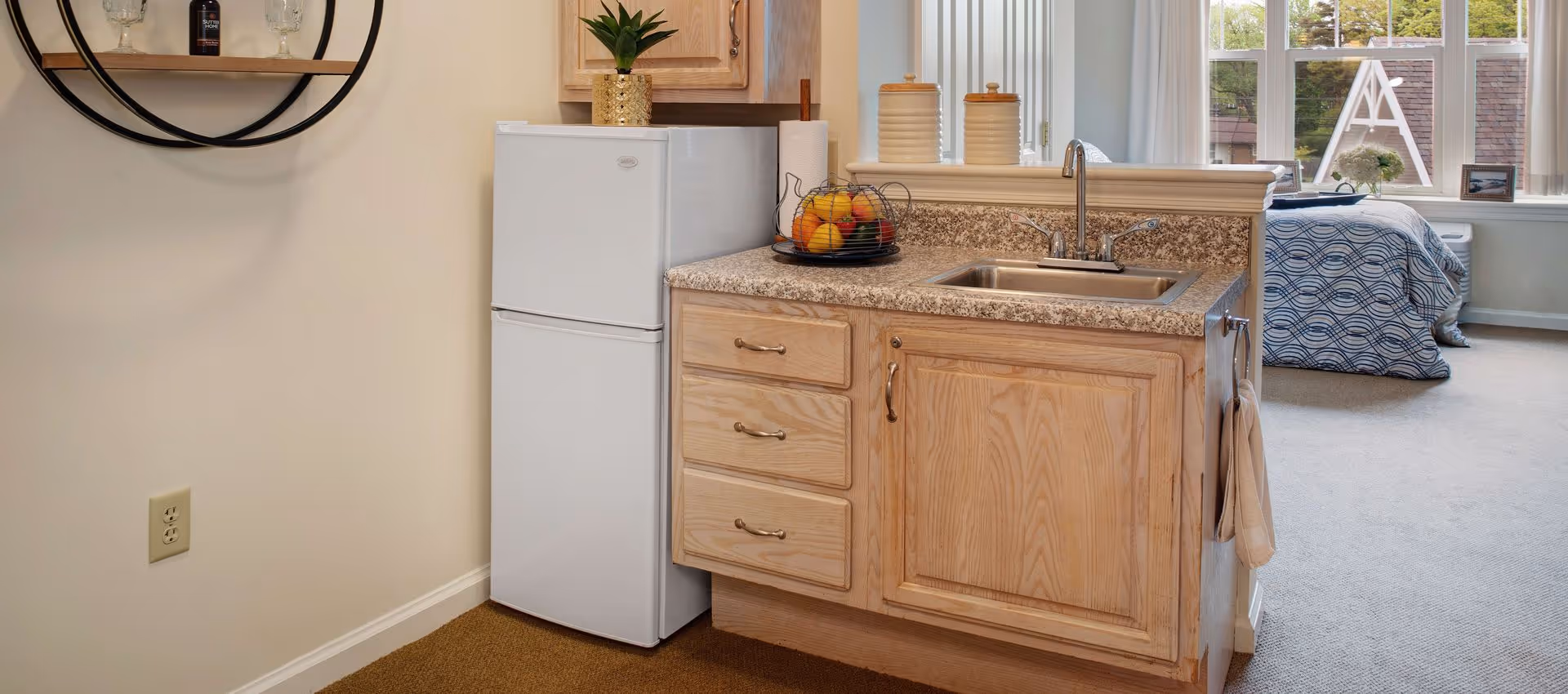Compact kitchenette with a mini refrigerator, sink and wooden cabinets opening into a studio bedroom.