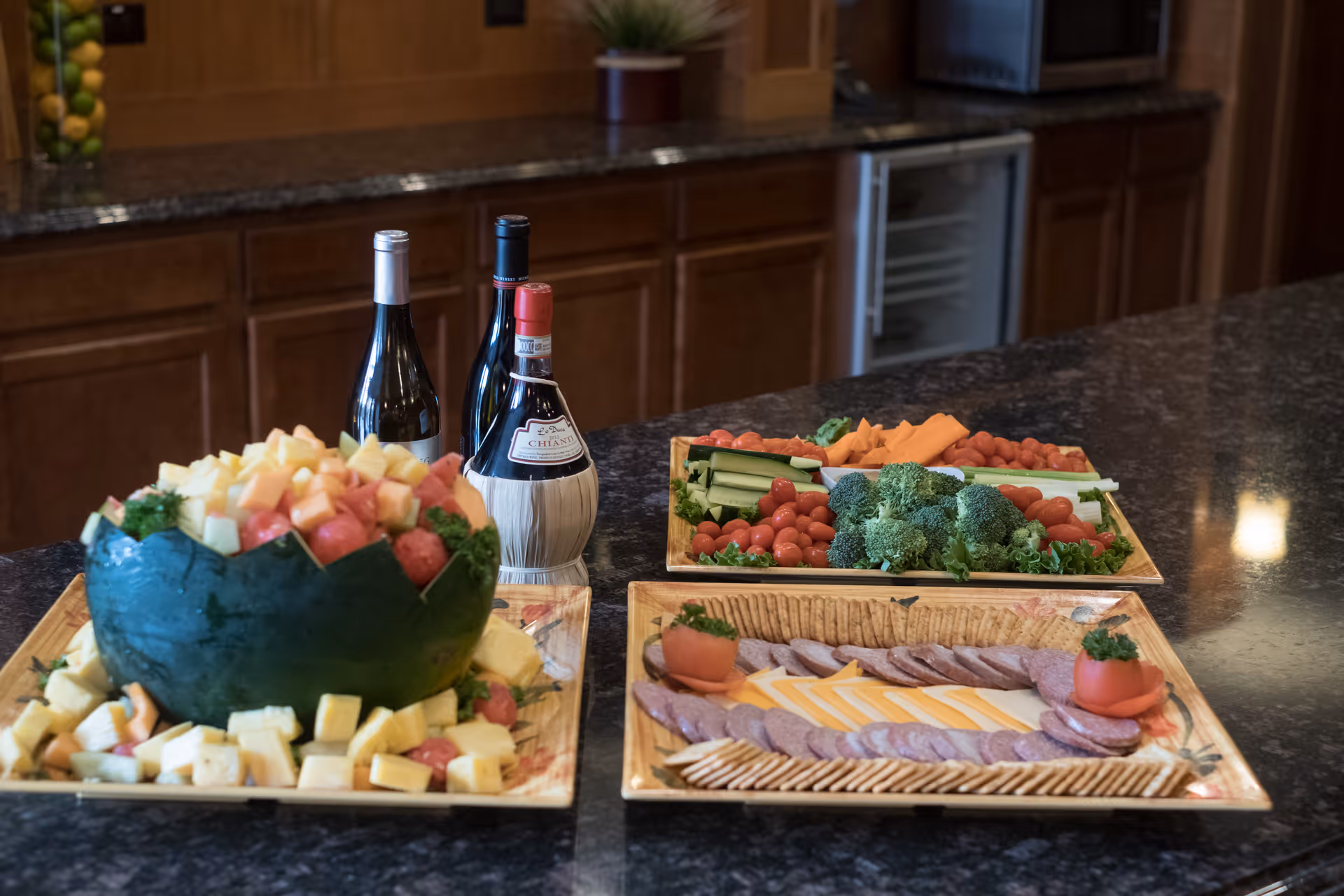A kitchen counter with three trays of food including a watermelon bowl filled with assorted fruit, a tray of sliced meats, cheese, and crackers, and a tray of fresh vegetables such as broccoli, cherry tomatoes, carrots, and celery. Three bottles of wine are placed behind the trays on the counter, with wooden cabinets and a small refrigerator in the background.