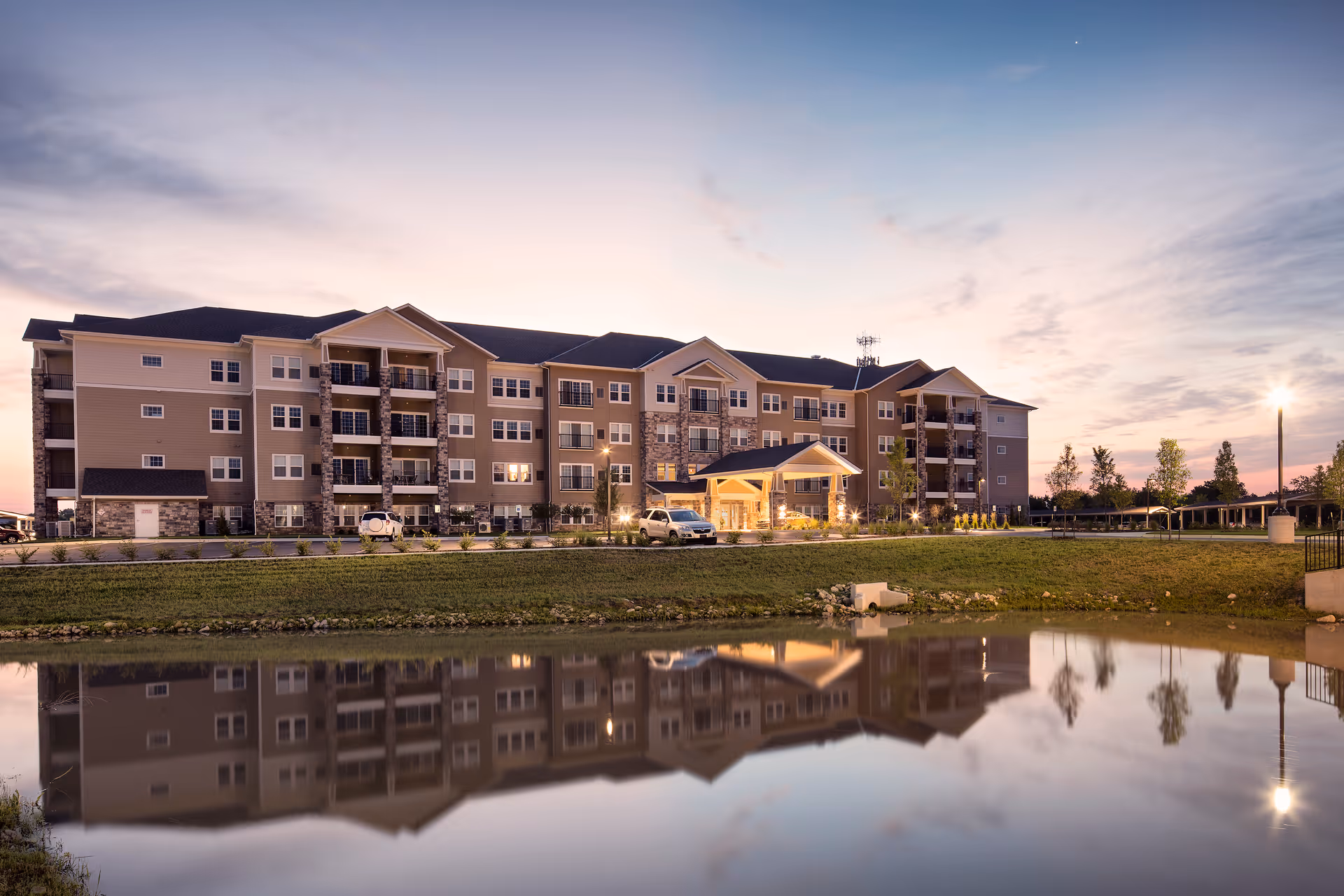 Four-story senior living apartment building at dusk with exterior lights on, reflected in a pond in the foreground.