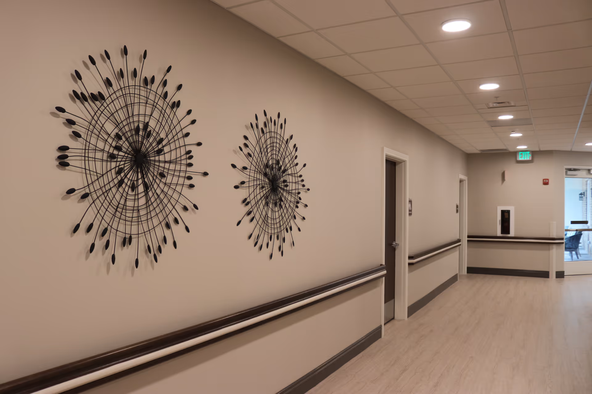 A clean, well-lit hallway in a senior living facility with beige walls, light wood flooring, and ceiling lights. The hallway features two decorative black metal wall art pieces resembling abstract sunbursts. There are handrails along the walls and several closed doors. At the end of the hallway, there is a glass door leading to another room with chairs visible inside.
