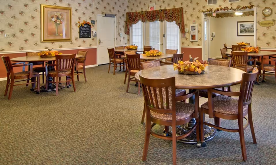A cozy dining room with several round tables and wooden chairs arranged neatly. Each table has a centerpiece with autumn-themed decorations. The walls are decorated with floral wallpaper, framed artwork, and a large mirror. There are windows with blinds and valances, and the carpet is patterned in muted tones.