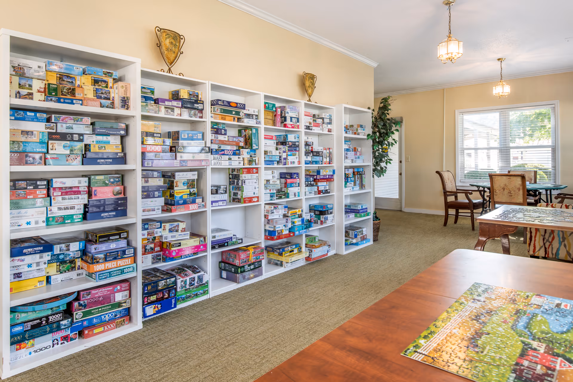 A bright room with beige walls and carpeted floor featuring multiple white shelves filled with numerous puzzle boxes. In the foreground, there are tables with partially completed jigsaw puzzles. The room has large windows letting in natural light and several chairs around the tables, creating a cozy activity space.