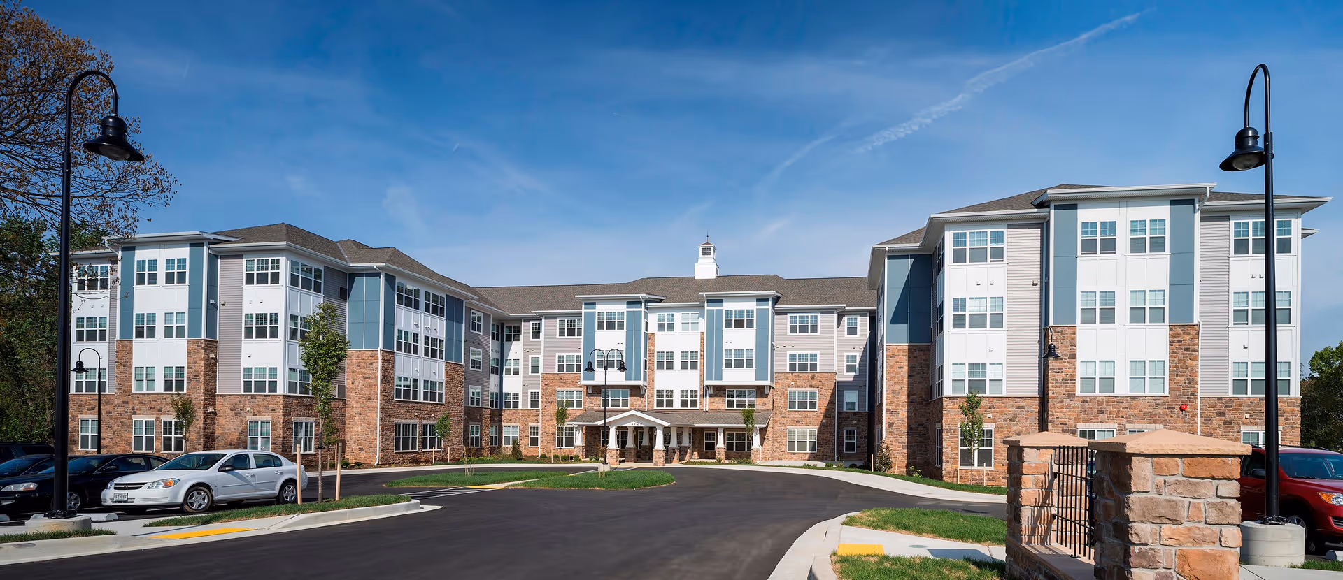 Front exterior view of a large, modern senior living facility named Greens at English Consul, featuring a multi-story building with a combination of brick and light-colored siding, numerous windows, a covered entrance, and a circular driveway with parked cars and street lamps under a clear blue sky.