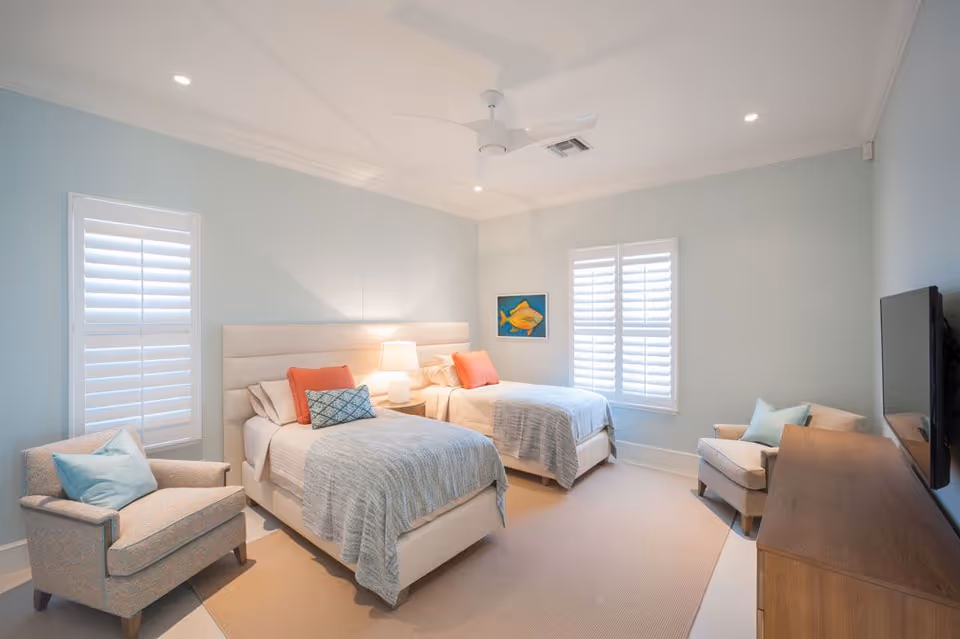 A bright and airy bedroom with two twin beds, each with light-colored bedding and coral accent pillows. Between the beds is a nightstand with a lamp. The room has light blue walls, two windows with white shutters, a ceiling fan, two armchairs with blue pillows, a wooden dresser, and a wall-mounted TV.