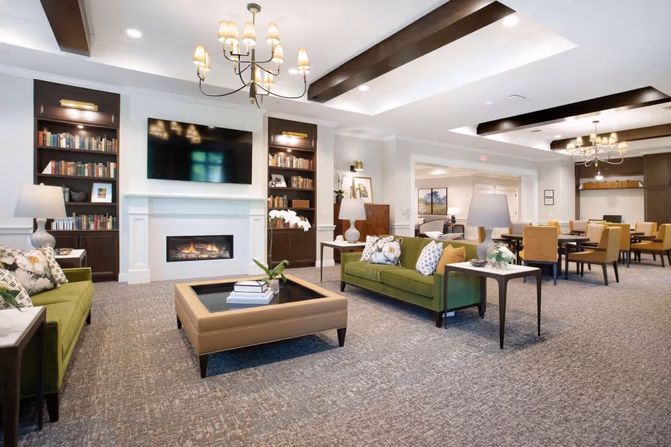 Spacious senior living common room with green sofas, a fireplace and wall-mounted TV, bookshelves, and dining tables in the background.