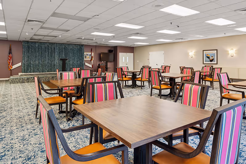 A spacious room with multiple wooden tables and chairs featuring colorful striped upholstery. The room has a patterned carpet, beige walls with framed artwork, and a small stage area with a podium and curtains. Two flags are positioned near the stage.