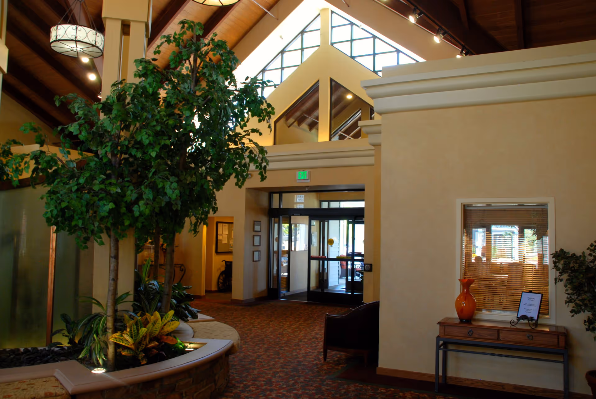 Interior view of a senior living facility lobby with a high vaulted ceiling, large windows, indoor plants, a seating area, and a wooden table with a decorative vase and framed sign near the entrance doors.