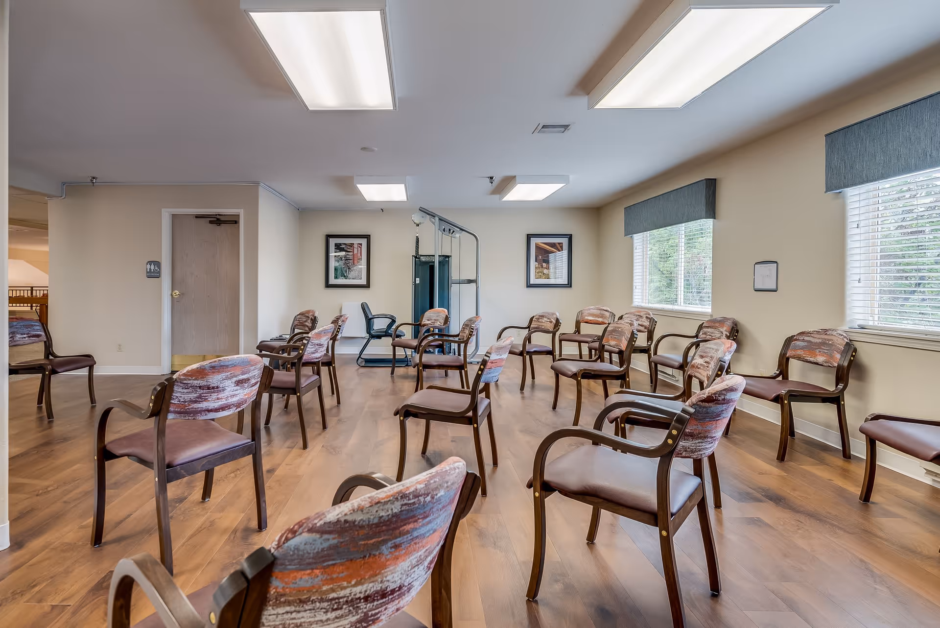 A spacious room with multiple wooden chairs arranged in a semi-circle on a wooden floor. The room has beige walls, two large windows with blinds and blue valances, fluorescent ceiling lights, two framed pictures on the far wall, and a piece of exercise equipment in the back center. A door with a restroom sign is visible on the left side of the room.