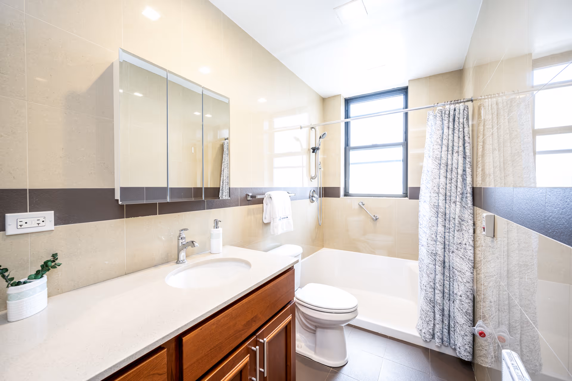 Bright and clean bathroom with a white bathtub and shower combination, a shower curtain, a toilet, a wooden vanity with a white countertop and sink, a wall-mounted mirror cabinet, a towel rack with a white towel, and a window letting in natural light.