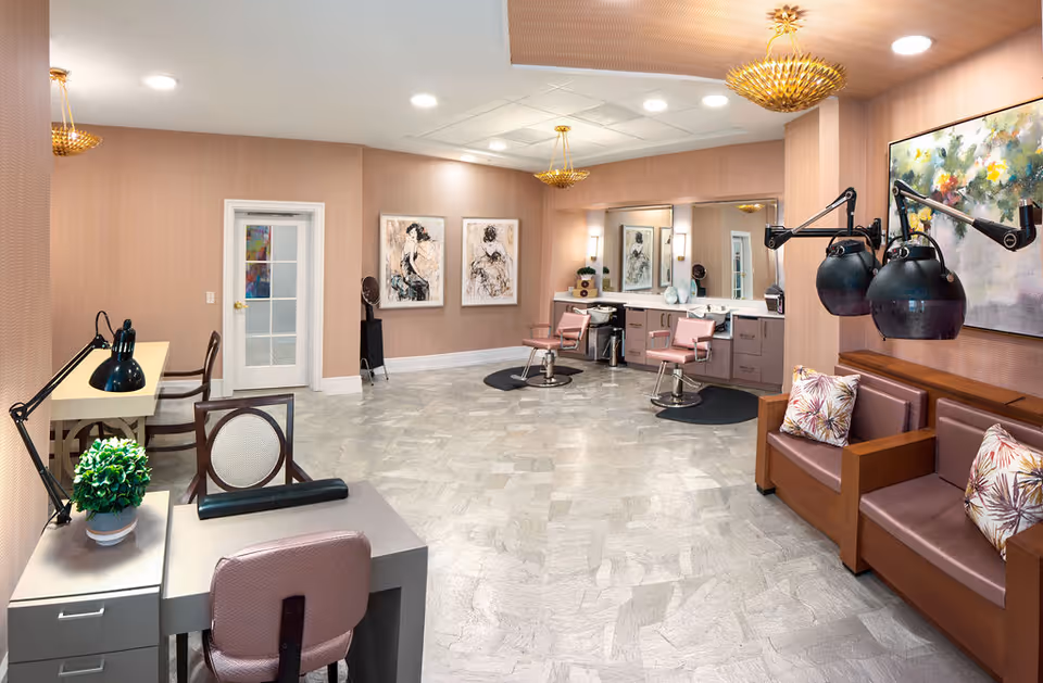 Interior view of a hair salon area in a senior living facility with two pink salon chairs in front of a large mirror, a seating area with brown cushioned chairs and hair dryers, a small desk with a lamp and plant, and framed artwork on the walls.